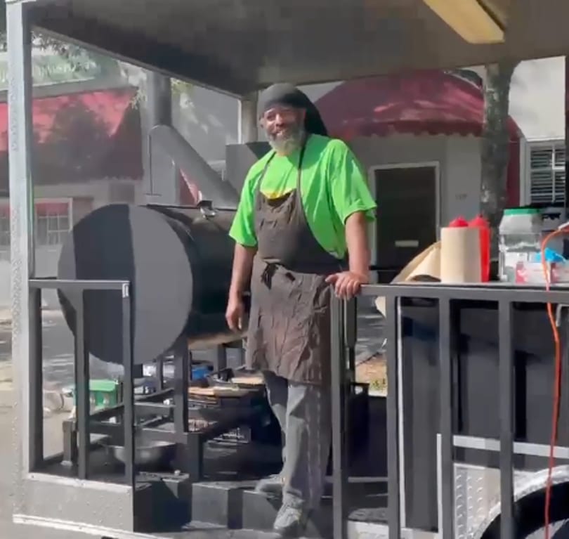 Man in a green shirt and apron standing next to a large black smoker on a food truck.