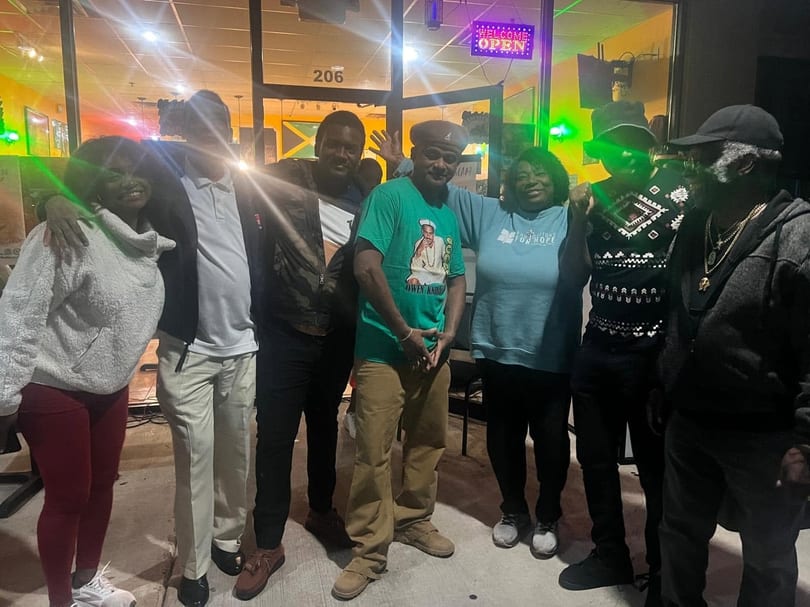 Seven people smiling and posing together outside a restaurant at night with an "OPEN" sign.
