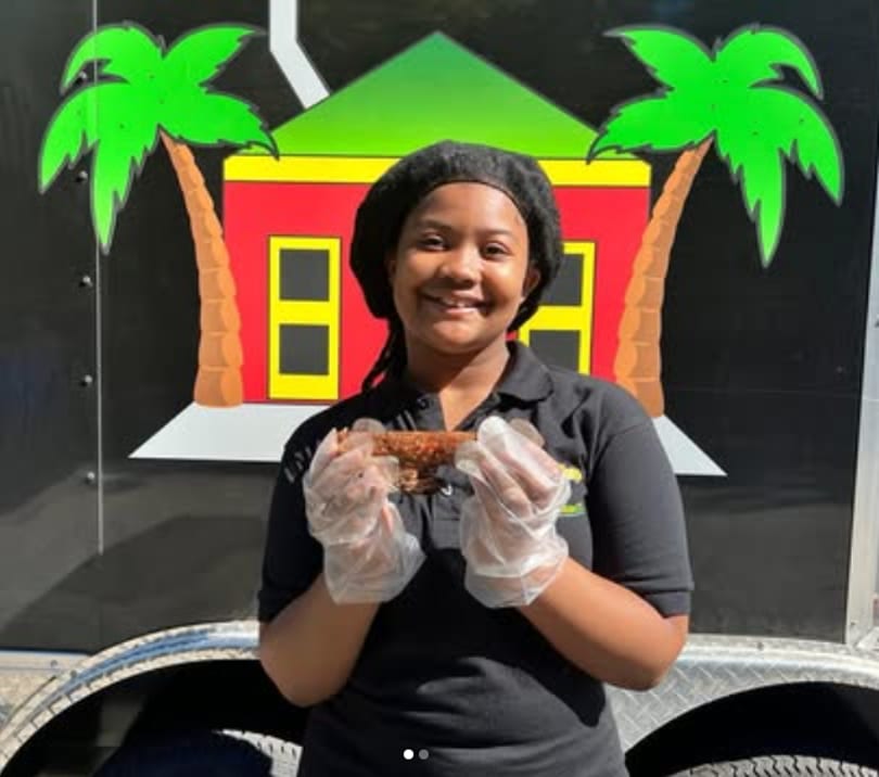 Smiling woman in gloves holding a piece of food in front of a food truck with a house logo.