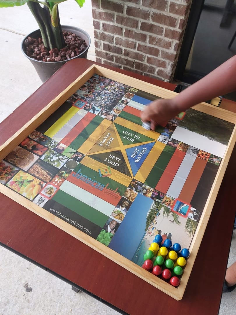 A person's hand plays Jamaican Ludo on a wooden board with colorful game pieces.