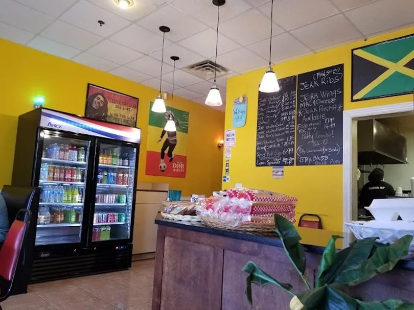 Interior of a vibrant Jamaican restaurant with yellow walls, a menu board, and a drink cooler.
