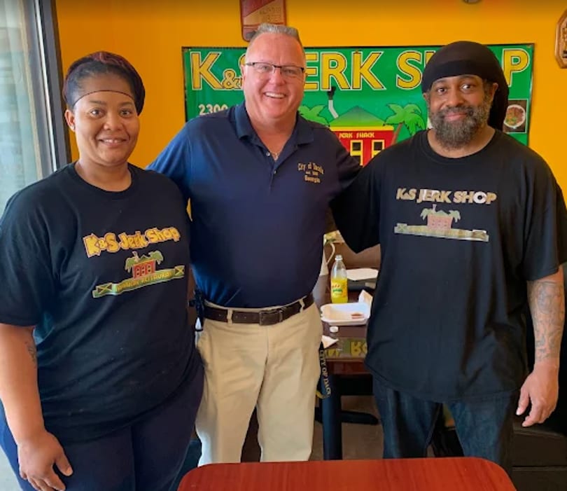 Three people, two wearing K&S Jerk Shop shirts, stand smiling in front of a colorful sign.