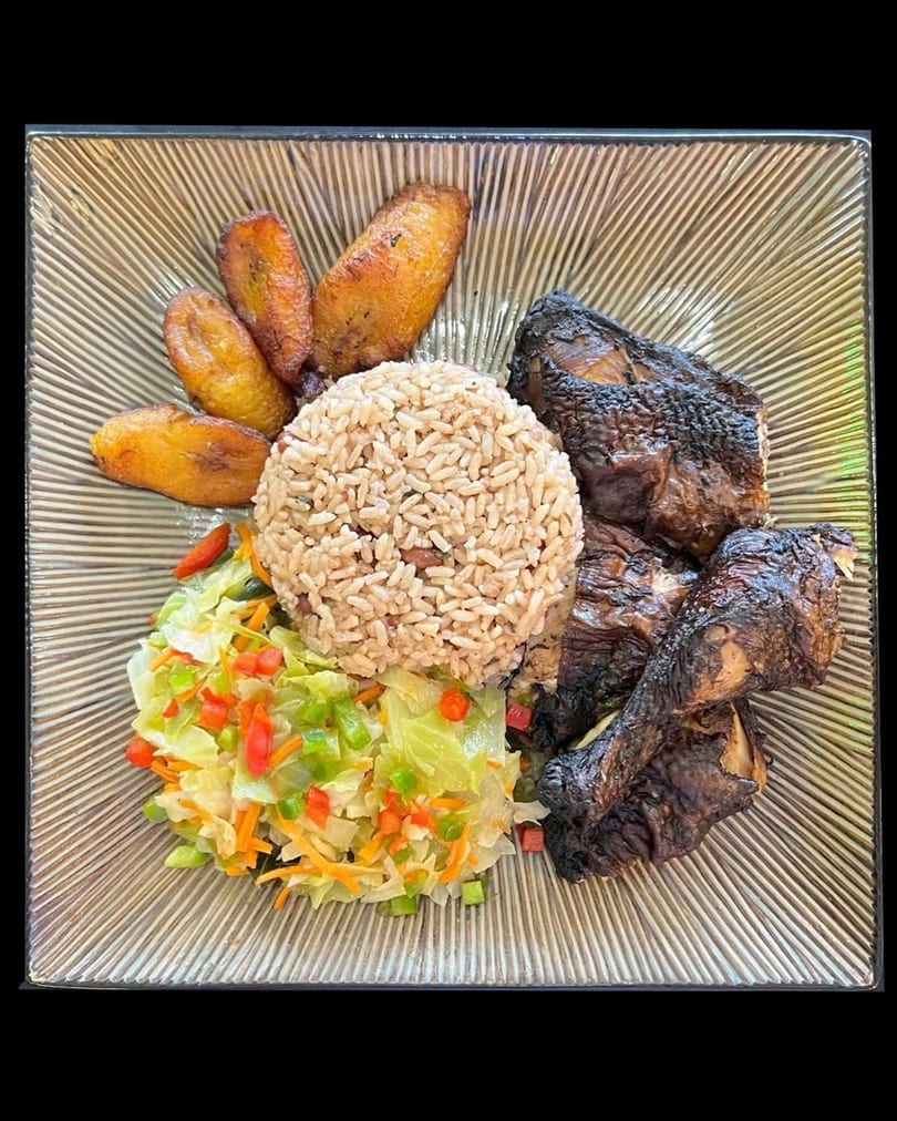 A plate of jerk chicken, rice and peas, fried plantains, and steamed cabbage with bell peppers.
