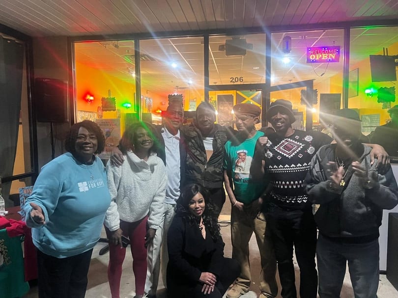 Group of nine people, including one woman kneeling, posing in front of a restaurant with colorful lights.