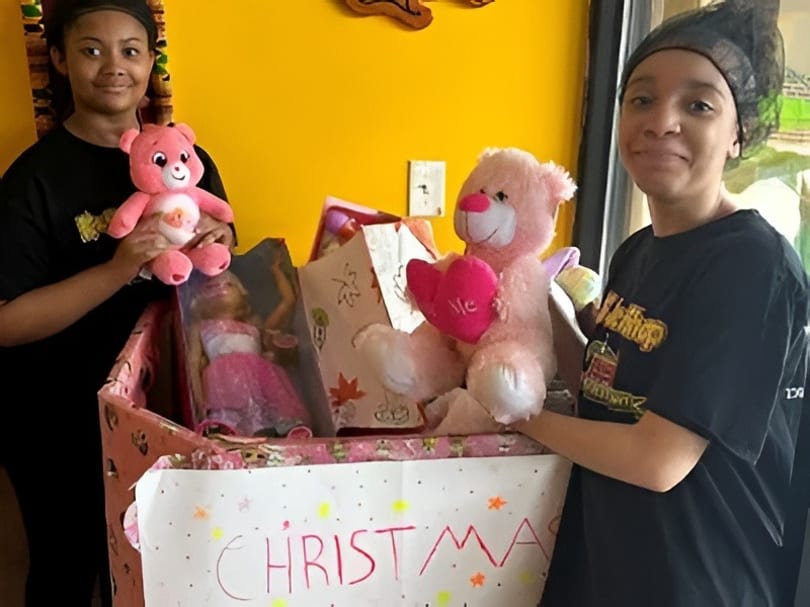 Two young women with a box of toys, including pink teddy bears and a doll, for Christmas.