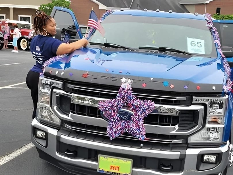 A woman decorates a blue Ford truck with patriotic garlands and an American flag.