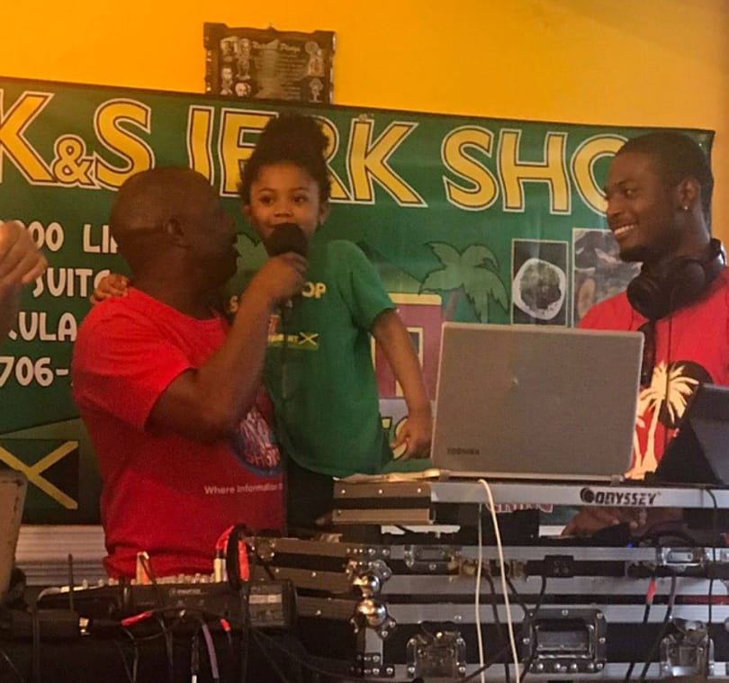 A man holds a child singing into a microphone, with a DJ smiling behind a laptop.