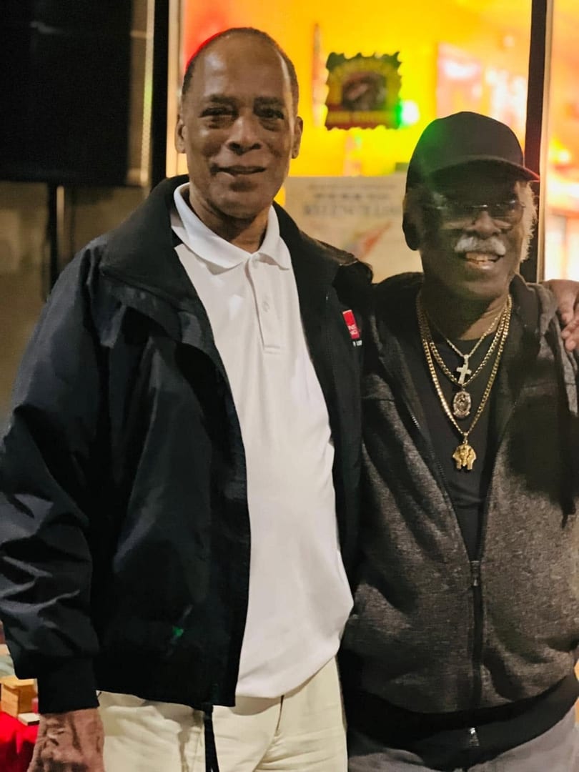 Two men smiling, one in a white polo and black jacket, the other in a black cap and gold chains.