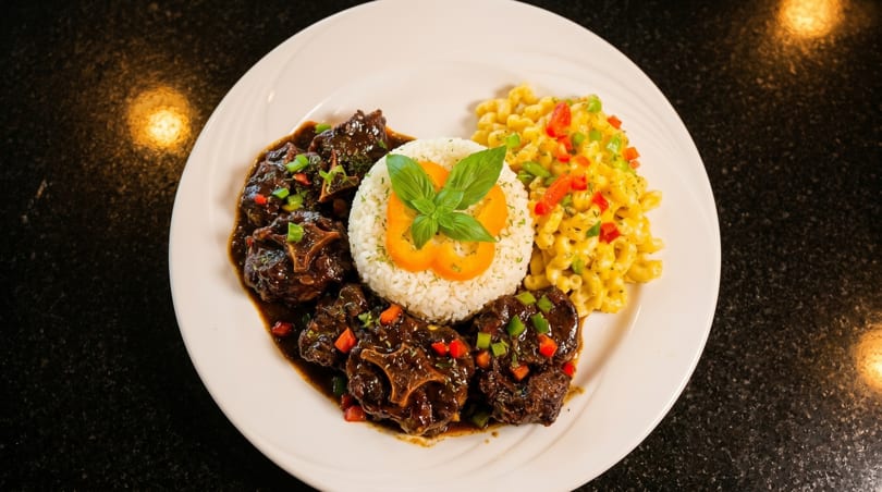 Oxtail stew with rice, mac and cheese, and bell peppers on a white plate.