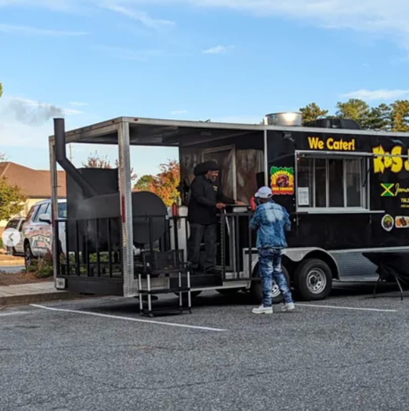 A black food truck with "We Cater!" and "K's Jamaican" text, serving a customer.