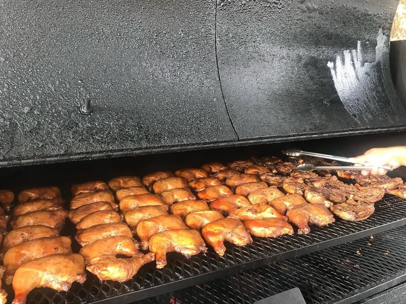 Rows of chicken quarters and other meat cooking on a large black grill, with tongs turning food.