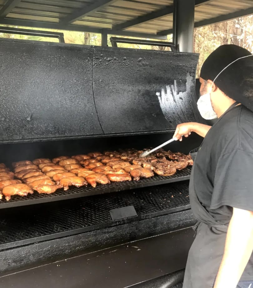Chef in a face mask grilling rows of chicken pieces on a large outdoor smoker.
