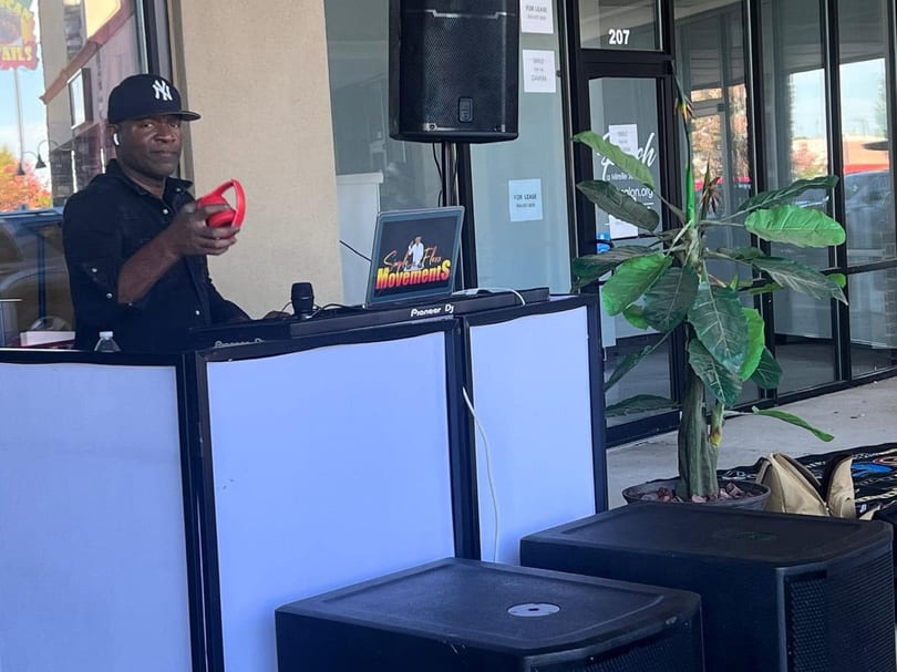 A DJ in a black shirt and Yankees cap holds red headphones, standing behind a Pioneer DJ setup.