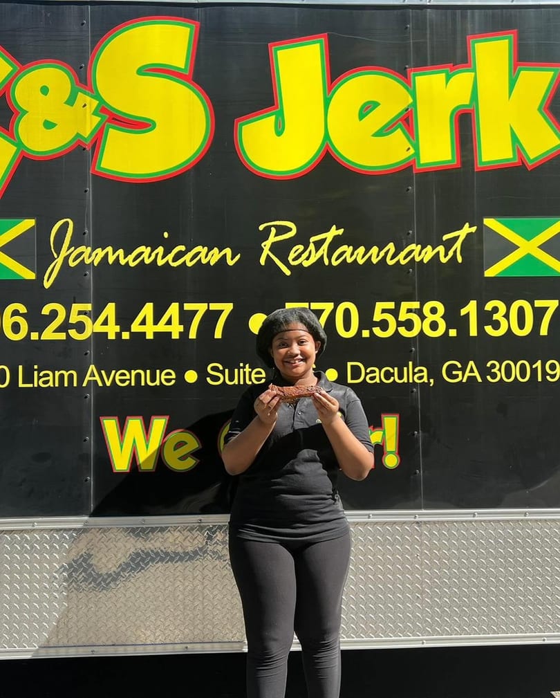 Smiling woman holding a piece of jerk chicken in front of the &S Jerk Jamaican Restaurant food truck.