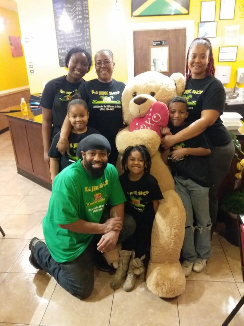Six people and a large teddy bear pose together in a restaurant with Jamaican decor.