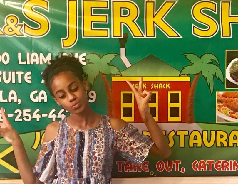 A young girl poses with peace signs in front of a green "Jerk Shack" restaurant banner.