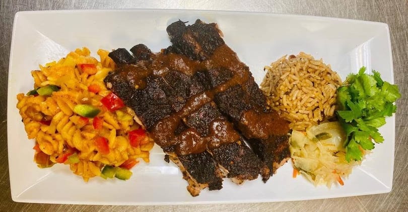 Grilled ribs with sauce, pasta salad, rice, and cabbage on a white rectangular plate.