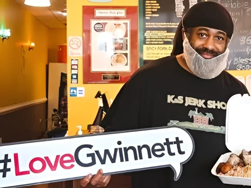 Man in a beard net and "K&S Jerk Shack" shirt holds a food container and #LoveGwinnett sign.