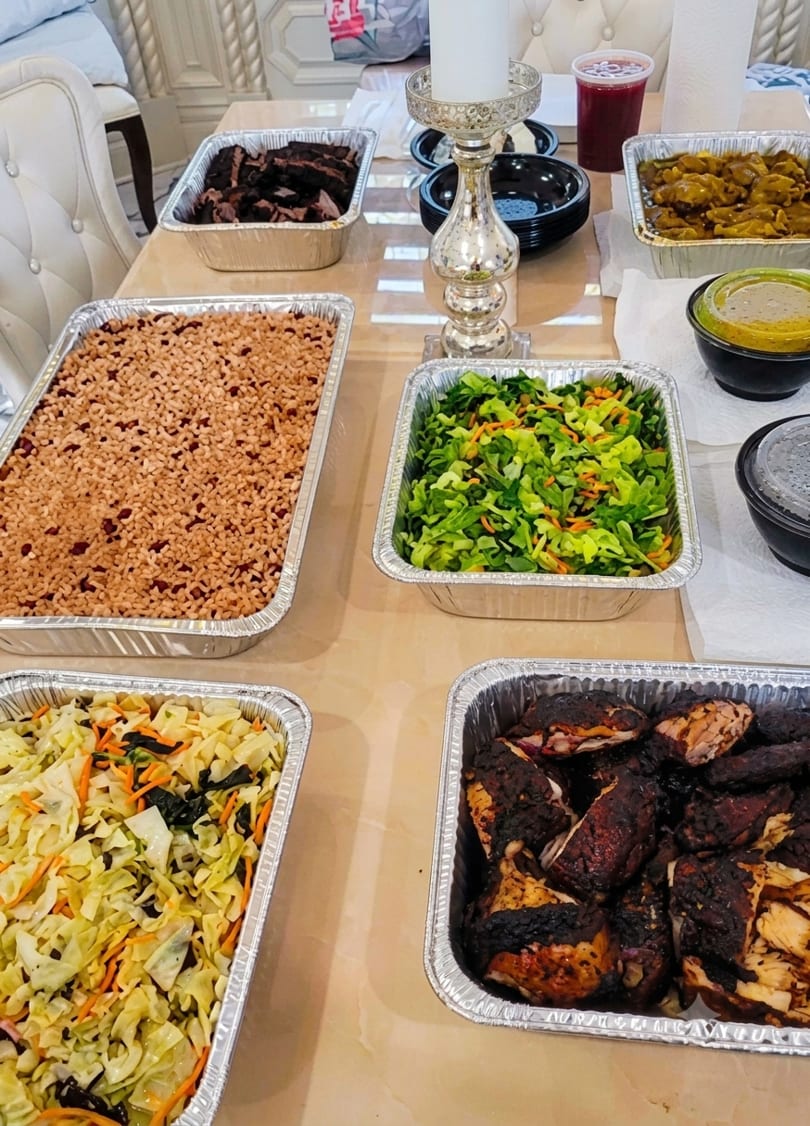 A buffet table laden with foil trays of rice and peas, jerk chicken, curry, and salads.