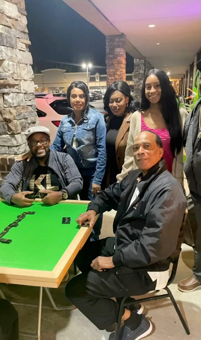 Group of five adults playing dominoes at a green table outdoors at night.