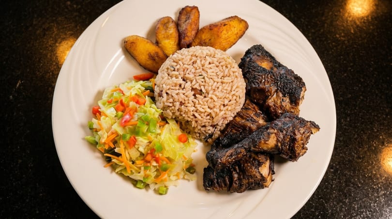 Jerk chicken with rice and peas, coleslaw, and fried plantains on a white plate.