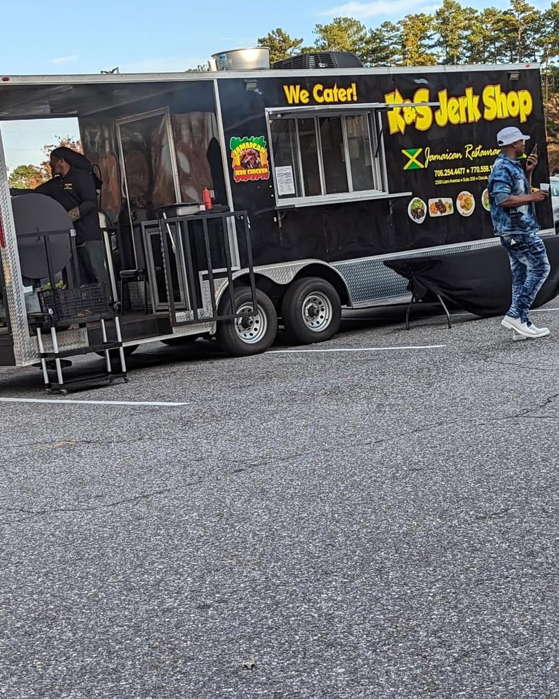 A black K&S Jerk Shop food truck with a person cooking and another walking by.