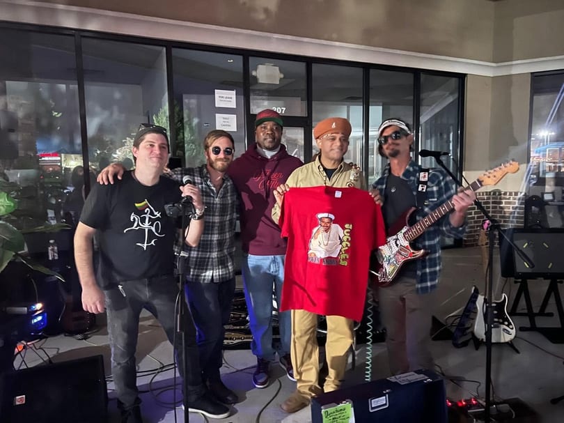 Five men, some holding instruments, pose with a red t-shirt featuring a man's portrait.